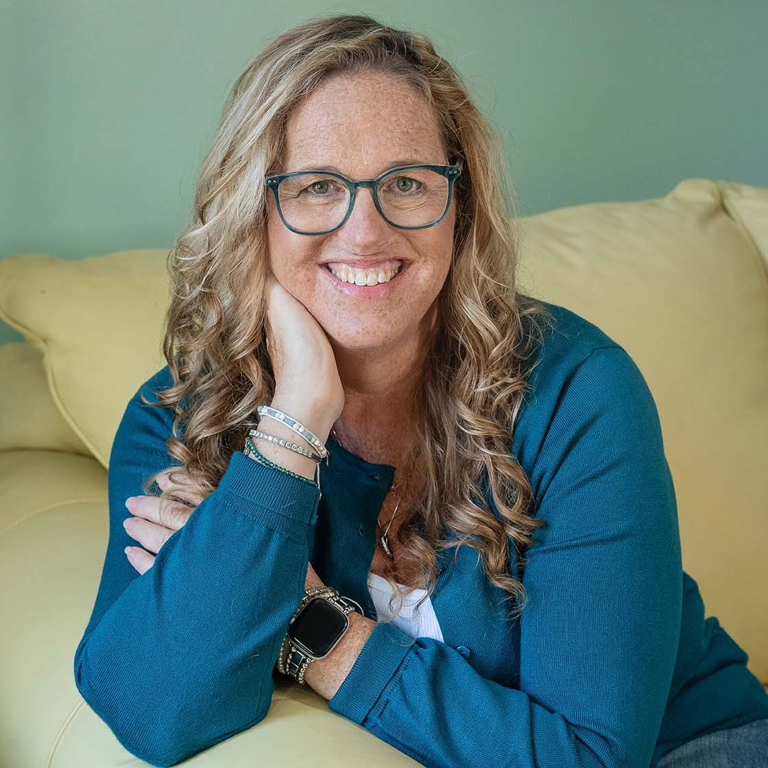 Jules McCullough with blonde hair and glasses sits on a light yellow leather couch, wearing a teal blue cardigan over a white top and blue jeans.