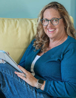 Jules McCullough with blonde hair and glasses sits on a yellow couch, holding a tablet. Working on children's book illustrations.